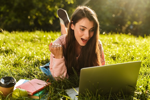 Foto piuttosto sorridente giovane studentessa che studia sul computer portatile mentre si trova su un prato al parco, celebrando