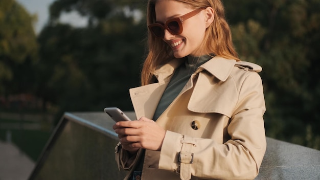 Bella studentessa vestita con trench e occhiali da sole sorridente in piedi sulla strada e in chat sullo smartphone