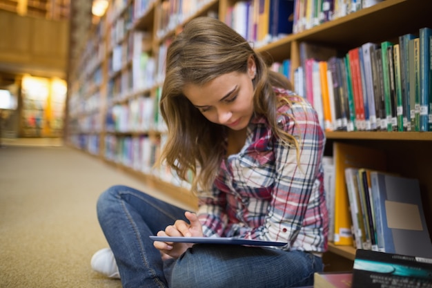 Studente grazioso che si siede sul pavimento delle biblioteche facendo uso della compressa