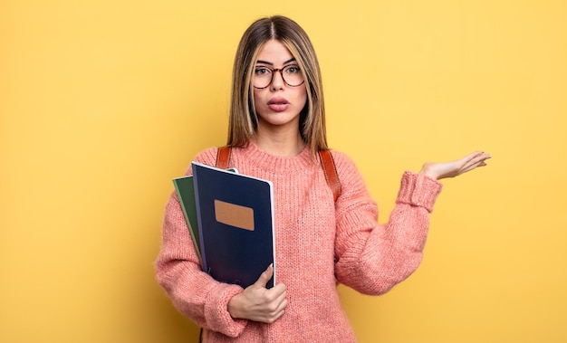 Pretty student woman looking surprised and shocked with jaw dropped holding an object books and backpack