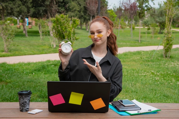 Pretty student working in park showing watch looking at camera