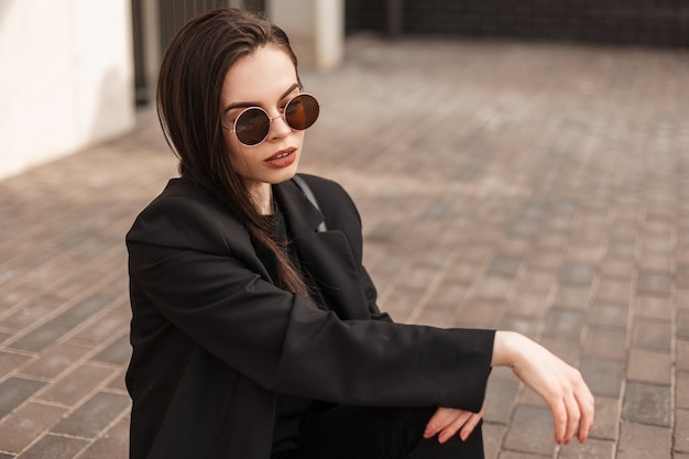 Pretty stylish young woman in fashionable youth black clothes in sunglasses sit on vintage tile in city. American girl fashion model rest on stone road on street. Fashion youth new collection.