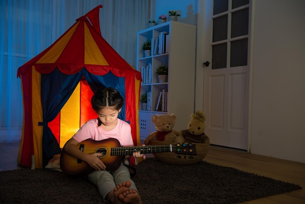 Foto bambina piuttosto dolce che tiene una piccola chitarra seduta davanti alla tenda dei bambini sul pavimento della camera da letto pratica l'ukulele durante la notte di vacanza con il suo giocattolo d'amore.