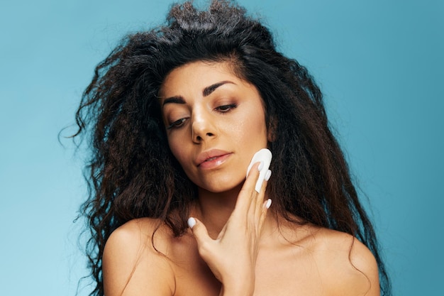 Pretty tanned latin woman cleaning face touching cheek cleaning\
facial skin looking down banner beautiful attractive curly lady\
using toner isolated on blue wall copy space close up portrait