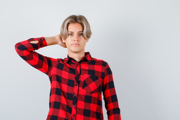 Ragazzo abbastanza teenager in camicia a quadri con la mano dietro la testa e guardando pensieroso, vista frontale.