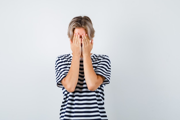 Pretty teen boy covering face with hands in striped t-shirt and looking scared. front view.