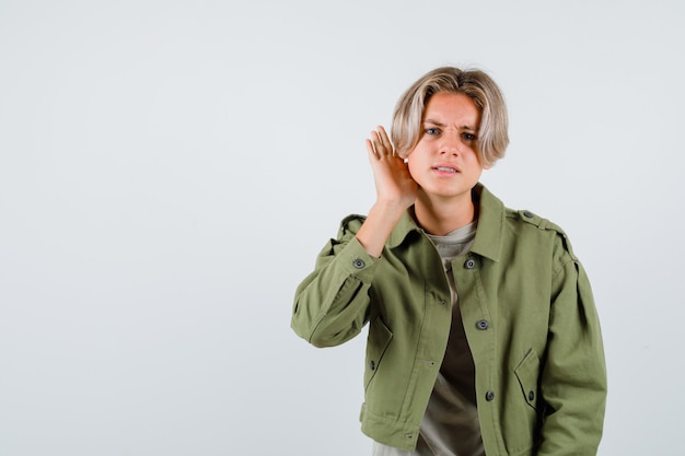 Photo pretty teen boy in green jacket keeping hand behind ear and looking perplexed