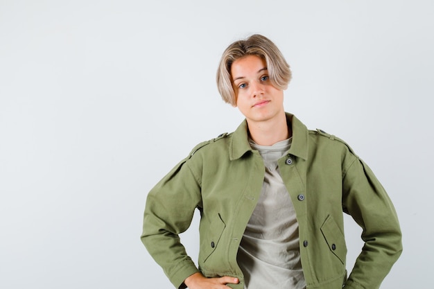 Pretty teen boy in green jacket keeping hands on waist and looking confident , front view.