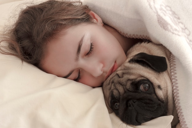 침대에서 퍼그 강아지를 안고있는 예쁜 십대 소녀 잔다
