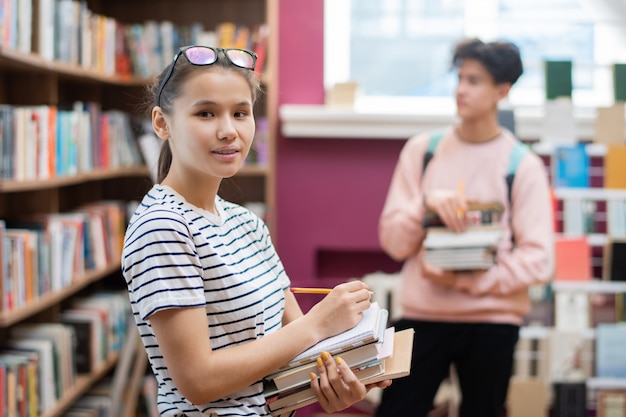 Pretty teenager with stack of books and pencil looking at you while standing in college library with classmate