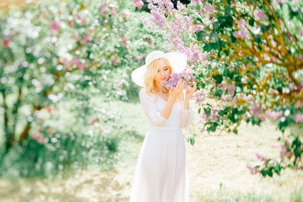 Pretty and tender young woman in white elegant dress and hat posing in blooming park.