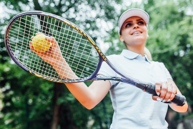 Pretty tennis player smiling at camera on a sunny day with racket on her hands.