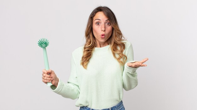 Pretty thin woman amazed, shocked and astonished with an unbelievable surprise and holding a washing dish brush