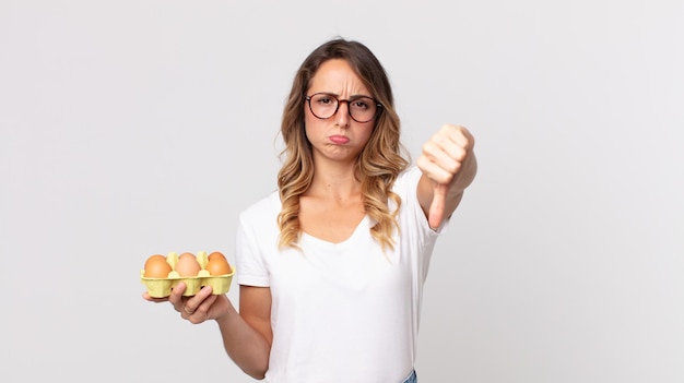 Pretty thin woman feeling cross,showing thumbs down and holding an eggs box