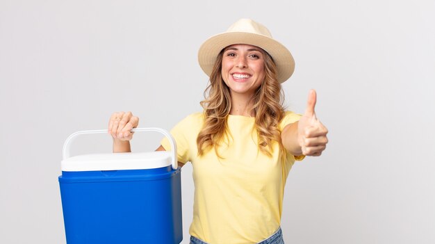 Pretty thin woman feeling proud,smiling positively with thumbs up and holding a  summer picnic fridge