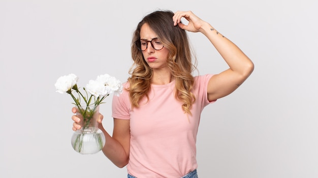 Pretty thin woman feeling puzzled and confused, scratching head and holding decorative flowers