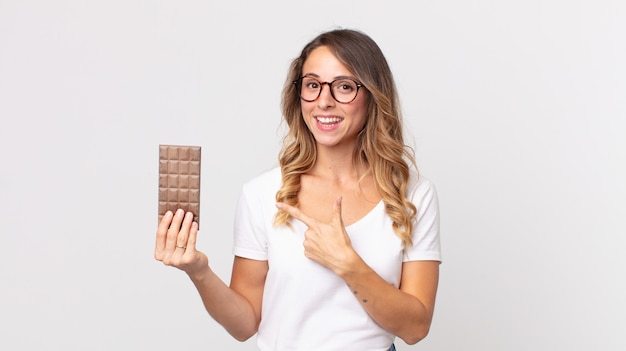Pretty thin woman looking excited and surprised pointing to the side and holding a chocolate bar