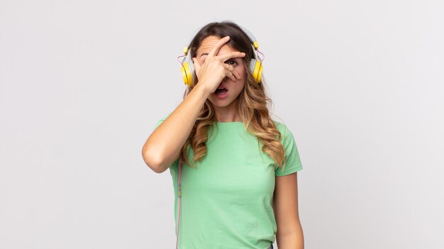 Pretty thin woman looking shocked, scared or terrified, covering face with hand listening music with headphones