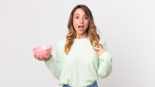 Pretty thin woman looking shocked and surprised with mouth wide open, pointing to self and holding an empty pot bowl