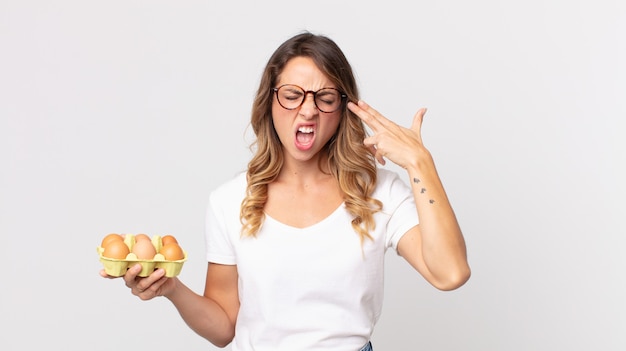 Pretty thin woman looking unhappy and stressed, suicide gesture making gun sign and holding an eggs box