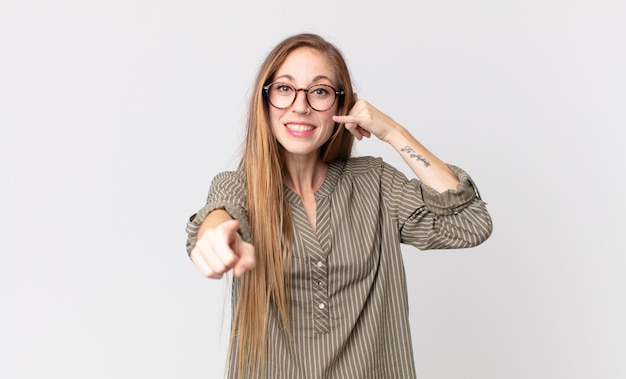 Pretty thin woman smiling cheerfully and pointing to camera while making a call you later gesture, talking on phone