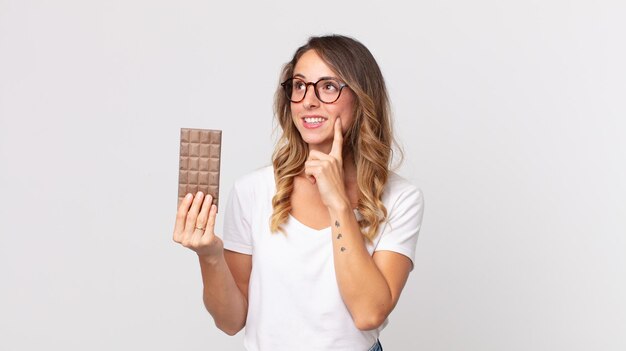 Pretty thin woman smiling happily and daydreaming or doubting and holding a chocolate bar