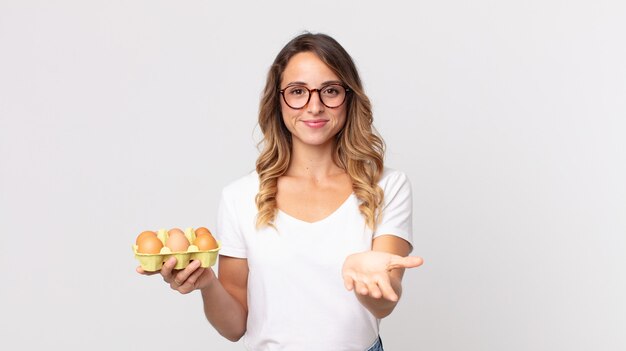 Donna piuttosto magra che sorride felicemente con amichevole e offre e mostra un concetto e tiene in mano una scatola di uova