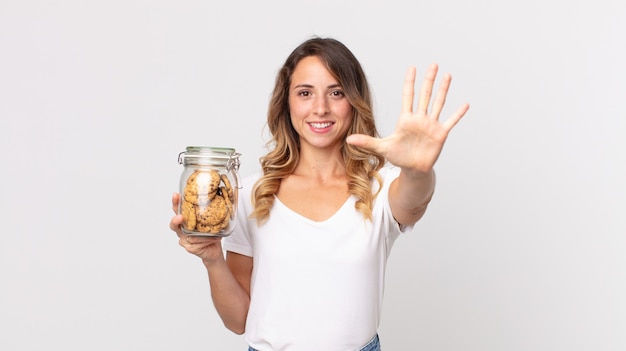 Pretty thin woman smiling and looking friendly, showing number five and holding a cookies glass bottle