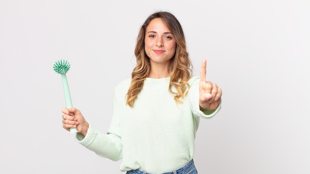 Pretty thin woman smiling and looking friendly, showing number one and holding a washing dish brush