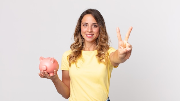 Pretty thin woman smiling and looking friendly, showing number two and holding a piggybank