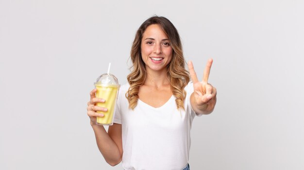 Pretty thin woman smiling and looking friendly, showing number two and holding a vanilla milkshake