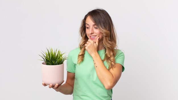 Pretty thin woman smiling with a happy, confident expression with hand on chin and holding a decorative plant