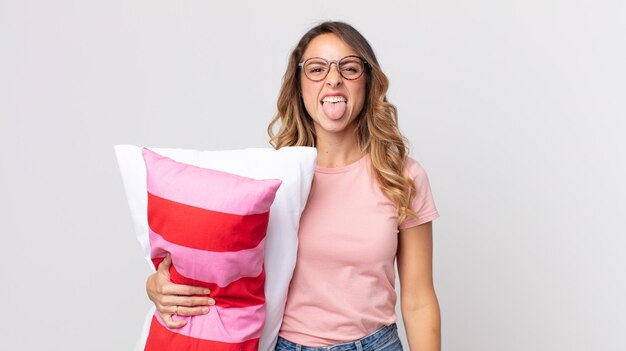 Pretty thin woman with cheerful and rebellious attitude, joking and sticking tongue out wearing pajamas and holding a pillow