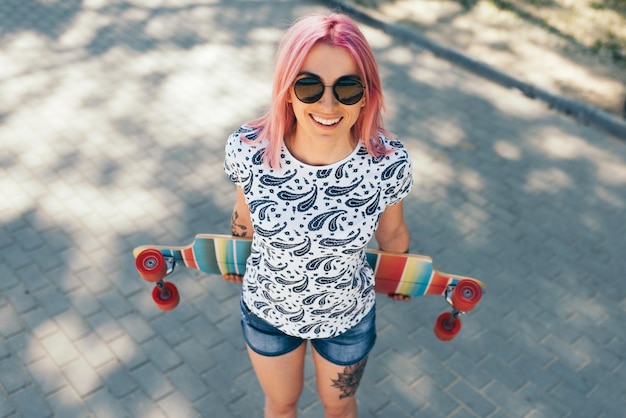 Bella vista dall'alto di una giovane donna attraente con i capelli rosa e con il longboard in strada indossando occhiali da sole guardando in alto bella ragazza sorridente che cammina con il longboard in strada