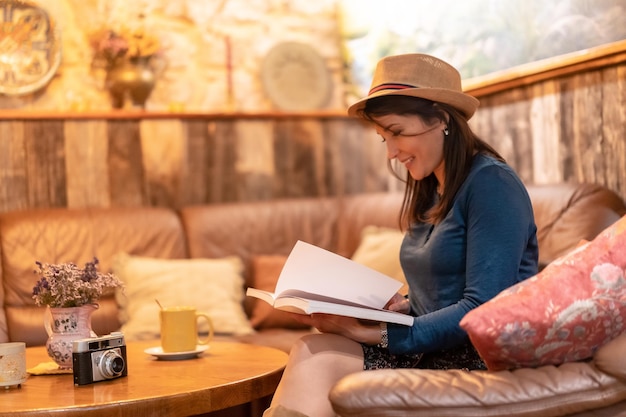 A pretty tourist with a hat and with a photo camera drinking tea in a cafe reading a book on a sofa inside the premises