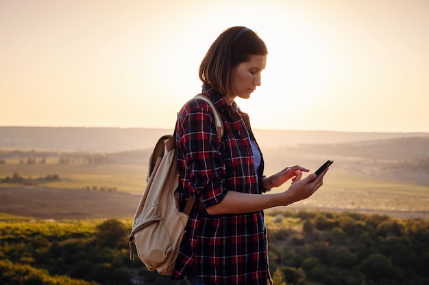 Симпатичная путешествующая женщина, стоящая на вершине горы на закате и пользующаяся мобильным телефоном