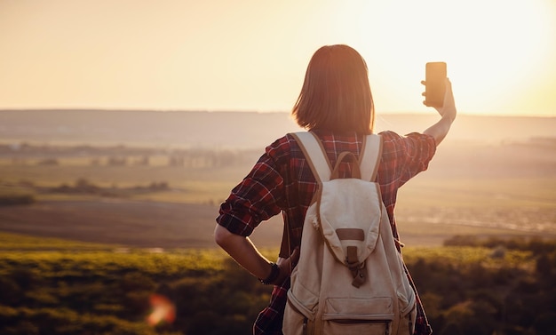 Pretty traveling woman standing on top of mountain at sunset and using mobile phone