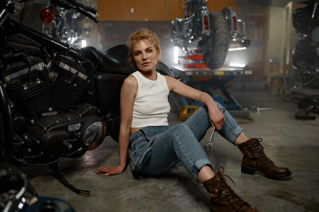 Pretty trendy girl mechanic sitting on garage floor nearby motorcycle