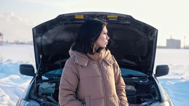 Pretty upset woman in warm jacket shrugs standing near broken automobile with open bonnet on snowy road in countryside on cold winter day