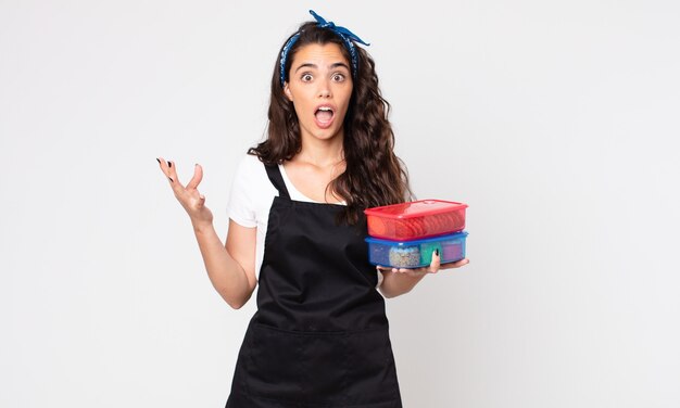 Симпатичная женщина удивлена, шокирована и удивлена невероятным сюрпризом и держит в руках посуду с едой.