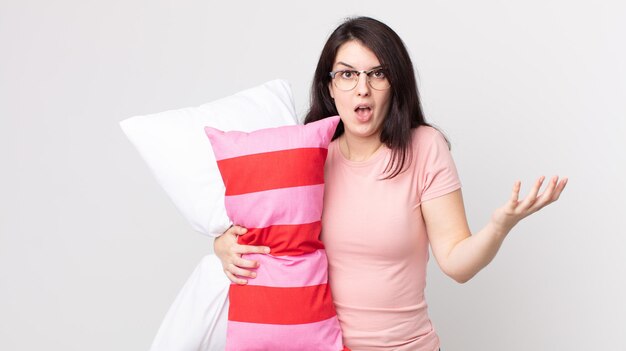 Pretty woman amazed, shocked and astonished with an unbelievable surprise wearing pajamas and holding a pillow