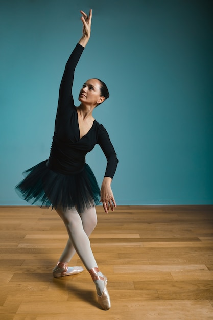 Pretty woman ballerina in tutu and pointe in black swimsuit posing in studio on blue background.