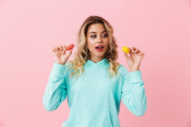 Pretty woman in basic clothing holding two macaron biscuits, isolated over pink wall