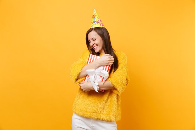 Pretty woman in birthday hat hugging with red box with gift, present celebrating, enjoying holiday isolated on bright yellow background. people sincere emotions, lifestyle concept. advertising area