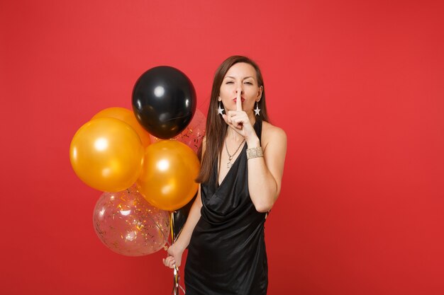 Pretty woman in black dress saying hush be quiet with finger on lips, shhh gesture celebrating, holding air balloons isolated on red background. Happy New Year birthday mockup holiday party concept.