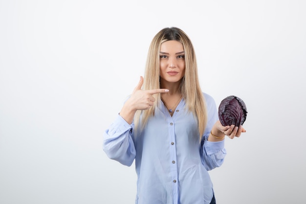 Pretty woman in blue outfit pointing at purple cabbage on white wall.