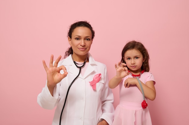 Pretty woman doctor in medical gown and a cute little girl, both with pink ribbon and phonendoscope around their neck, gesticulate with an OK sign to support breast cancer patients and survivors.