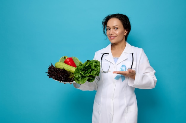 Pretty woman, doctor nutritionist in white medical gown with a blue awareness ribbon shows on plate full of healthy raw vegan eating. World Diabetes Day concept on colored background with space for ad