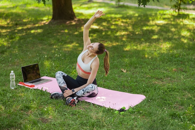 Pretty woman does side bends sitting near laptop in green summer park