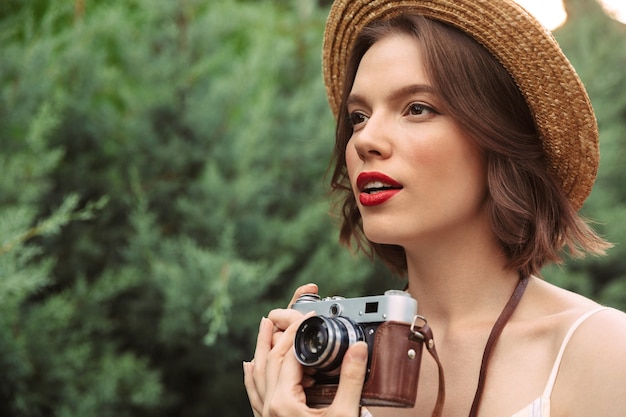 Bella donna in abito e cappello di paglia che fa foto sulla fotocamera retrò mentre guarda lontano all'aperto